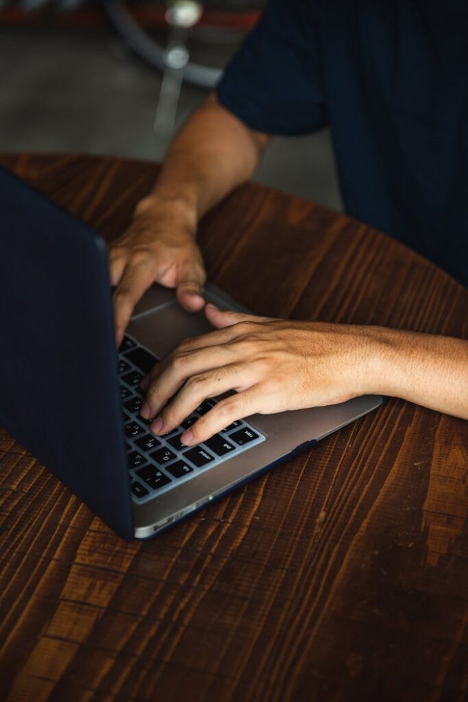 Close-up of a man typing on a laptop at a wooden table, indoor setting.
